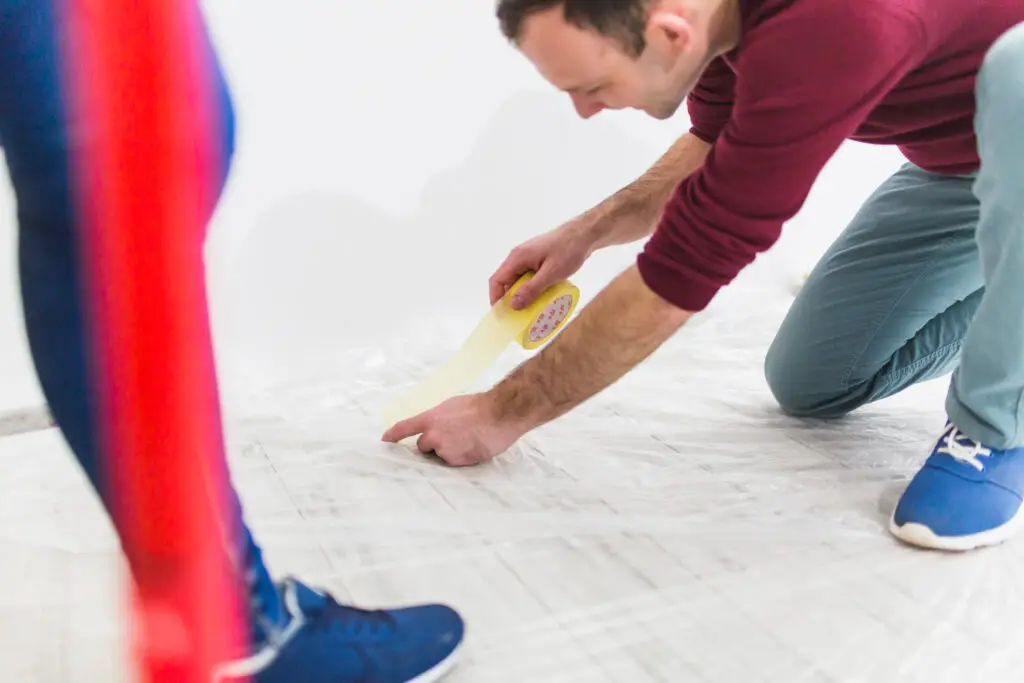 Two people lay tape on the floor; one person kneels with blue shoes, focused on precise placement and teamwork in a bright room.