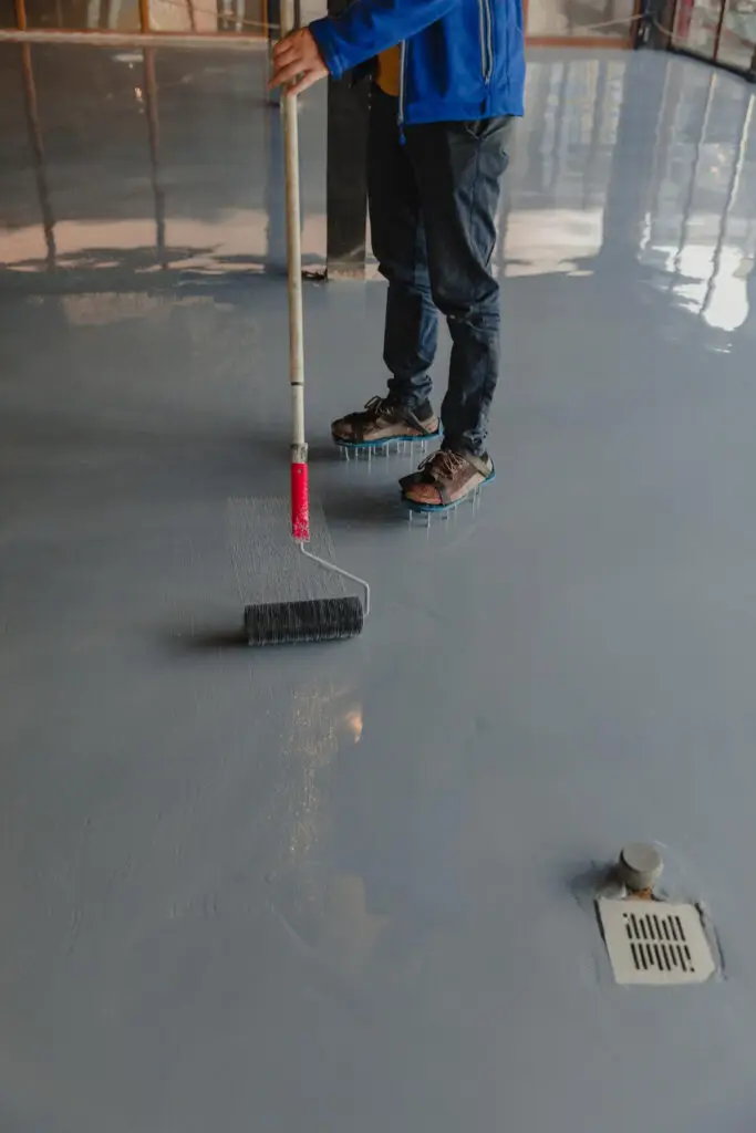 A person applies a resin coating on a floor using a roller, wearing spiked shoes for even distribution, in a construction setting.