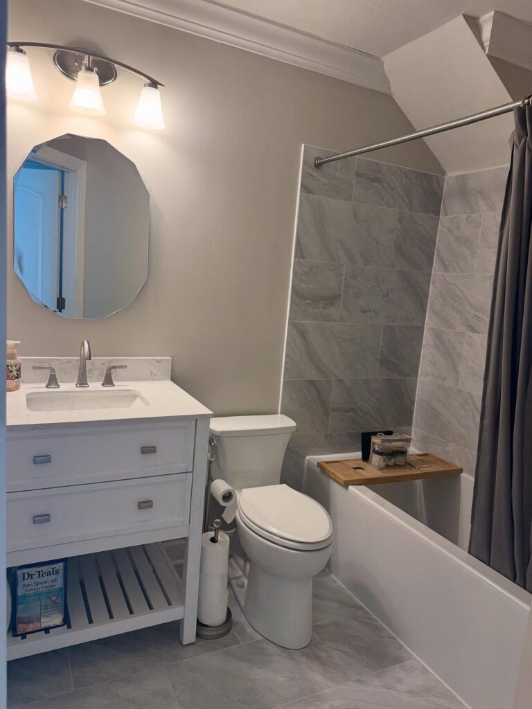 A modern bathroom features a sink, mirror, toilet, and bathtub with gray tiles and a wooden bath tray under soft lighting.