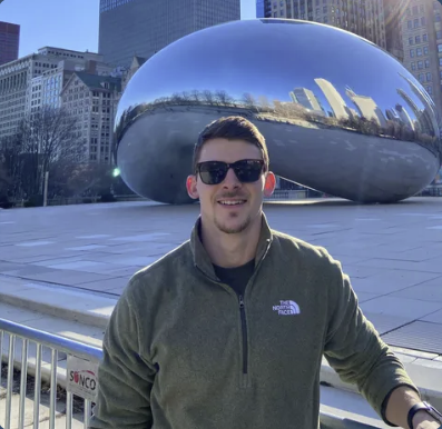 A person wearing sunglasses stands in front of Cloud Gate, also known as "The Bean," in Chicago’s Millennium Park, on a sunny day.