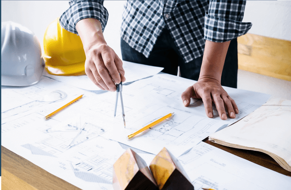 A person stands over architectural plans, using a compass. Two hard hats and pencils are on the table, suggesting design and construction work.