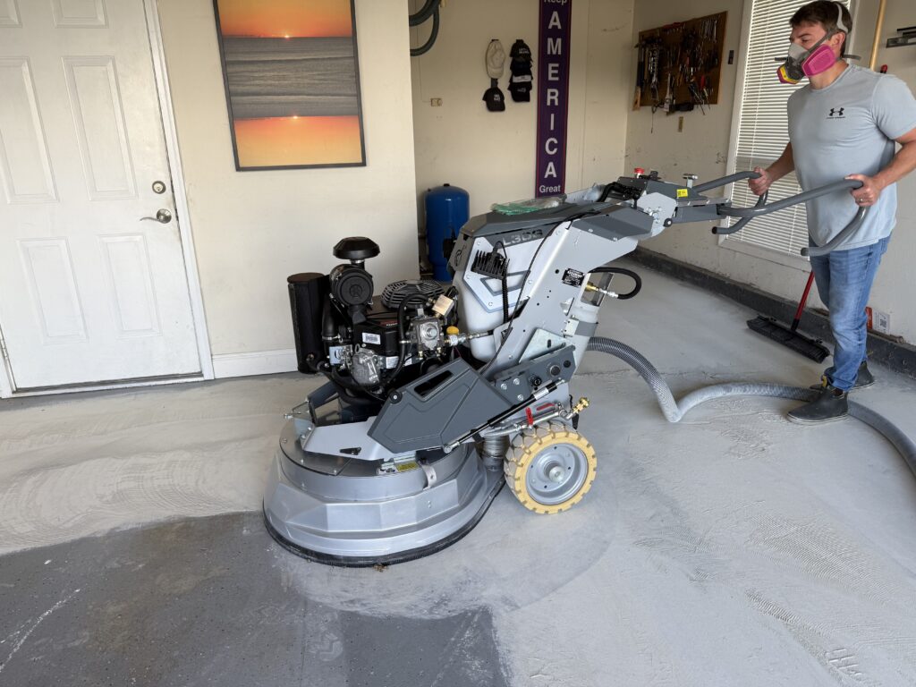 A person operating a floor grinder in a garage beside a wall with tools and a landscape painting.