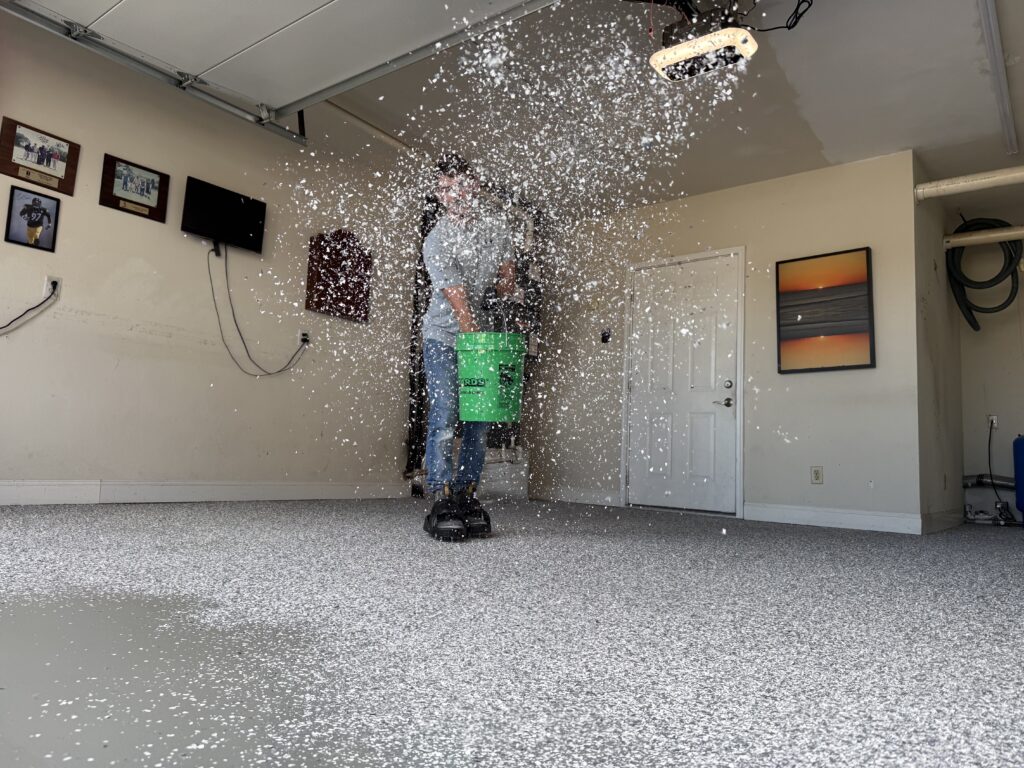 A person throws white material onto a floor in a garage. Walls have framed pictures and a television. Green bucket in use.