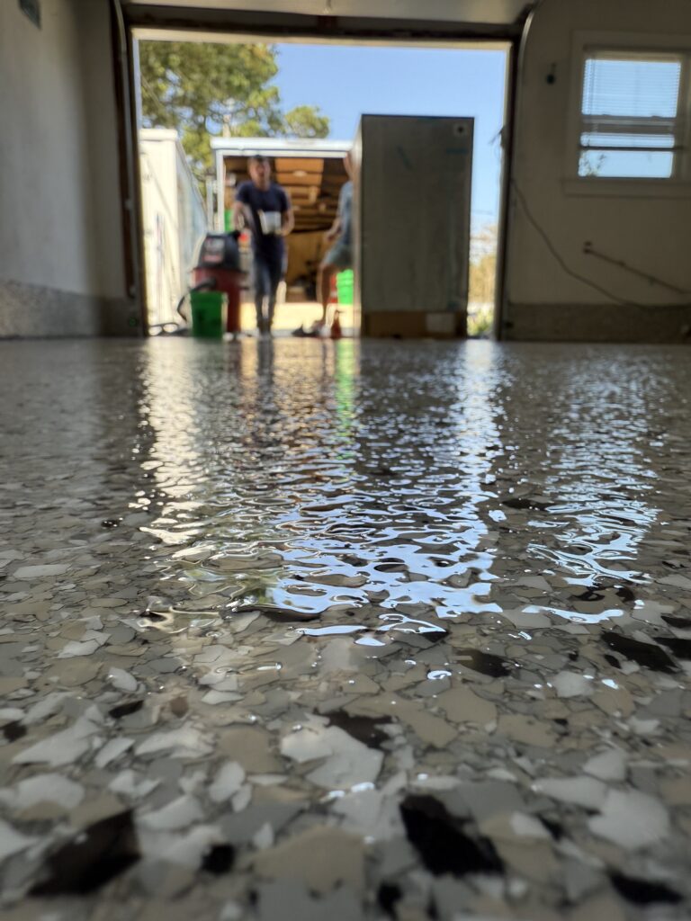 Shiny, speckled garage floor with reflections. Two people visible through a garage door, carrying cleaning supplies. Sunlight illuminates the scene from outside.