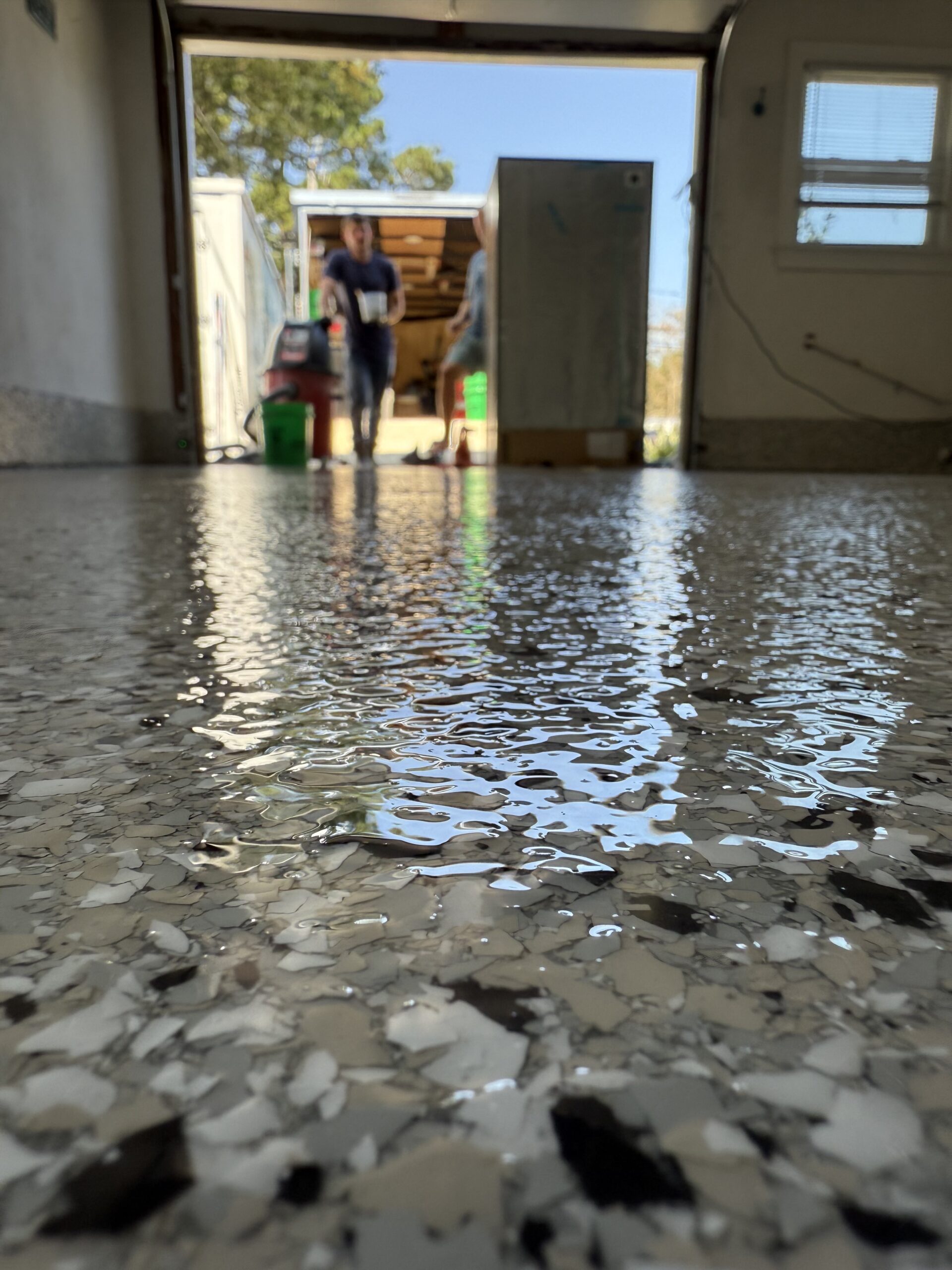 Shiny, speckled garage floor with reflections. Two people visible through a garage door, carrying cleaning supplies. Sunlight illuminates the scene from outside.